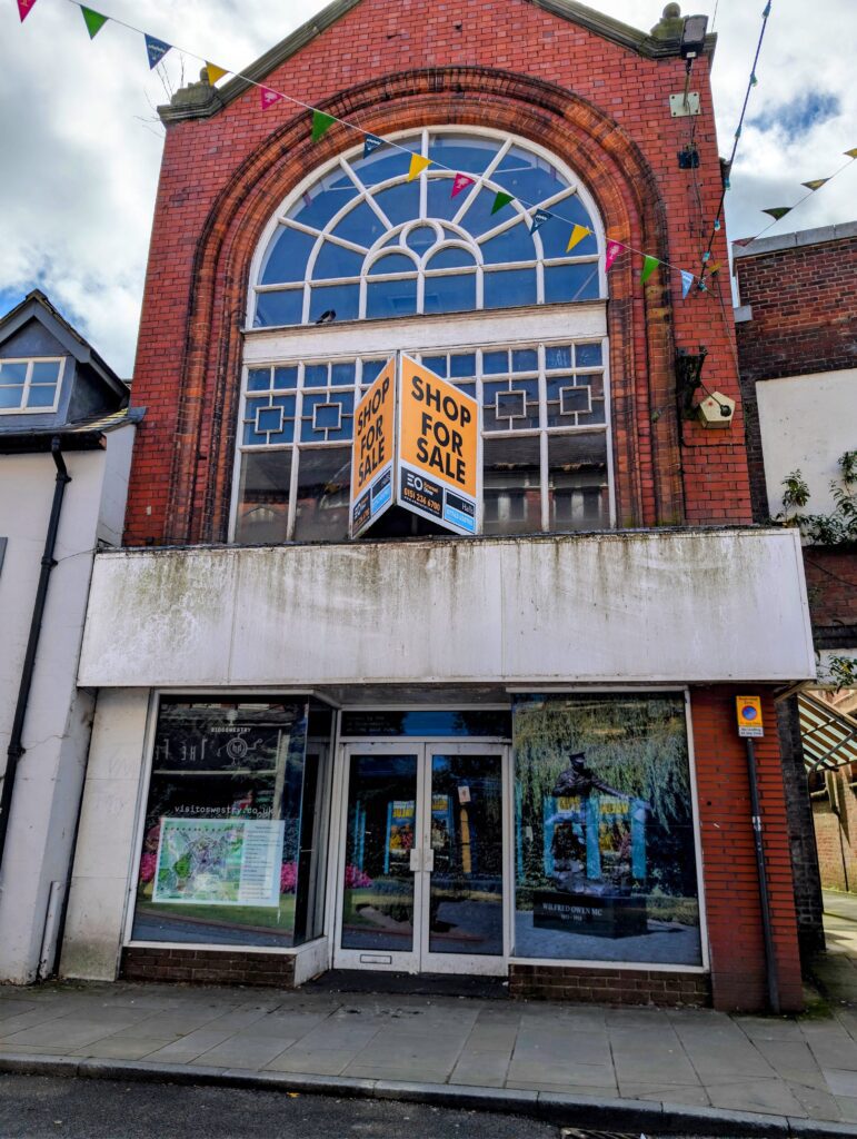 A straight-on exterior shot of a traditional red-brick building with a vacant ground-floor retail shopfront. The building features a large, arched multi-pane window on the upper floor, with a prominent orange and black "SHOP FOR SALE" sign fixed to the center window frame. Colourful overhead bunting stretches across the top of the frame against a blue sky with white clouds. The ground-floor windows and double glass doors are covered with large posters displaying local town information, map details, and an image of the Wilfred Owen statue.