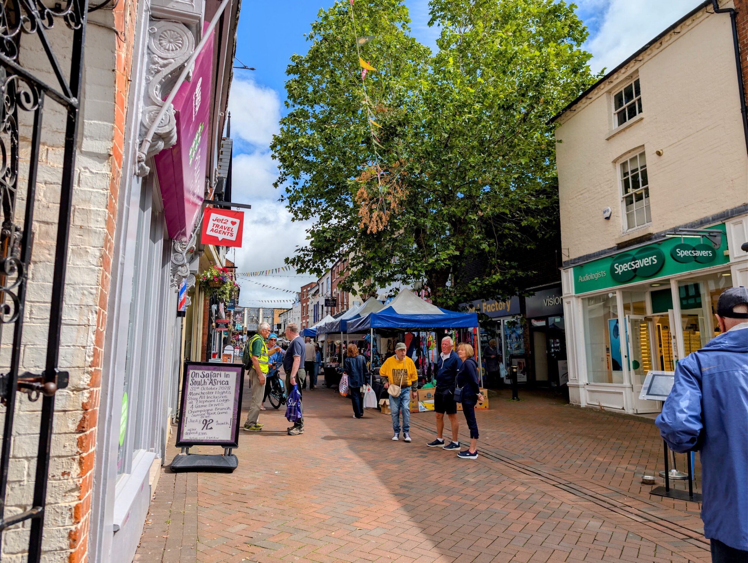 A wide shot looking down a brick-paved shopping street with a large tree in the centre. Market tents are set up along the path, positioned between established storefronts like Specsavers and a travel agency. People are scattered throughout the space, talking and shopping.