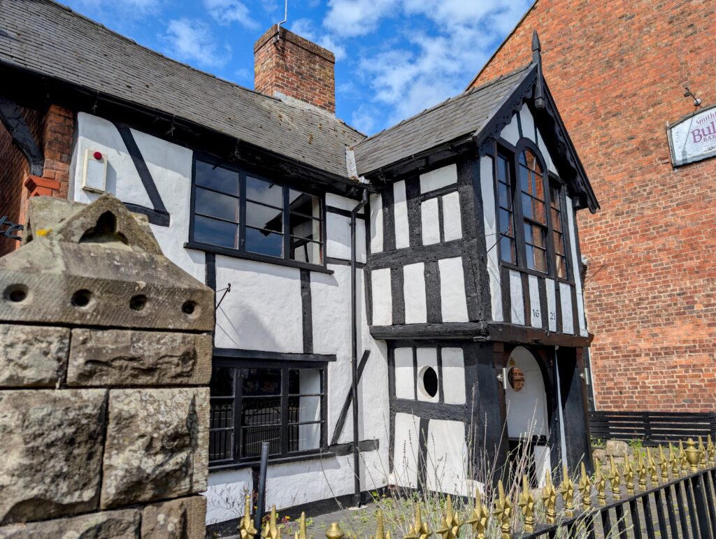 A historic, timber-framed building known as the Black Gate, featuring a classic white exterior with exposed black wooden beams. The two-story structure has multi-paned windows, a prominent gabled porch entryway with the year "1621" faintly visible on the woodwork, and a stone wall and metal railings in the foreground under a blue sky with light clouds.
