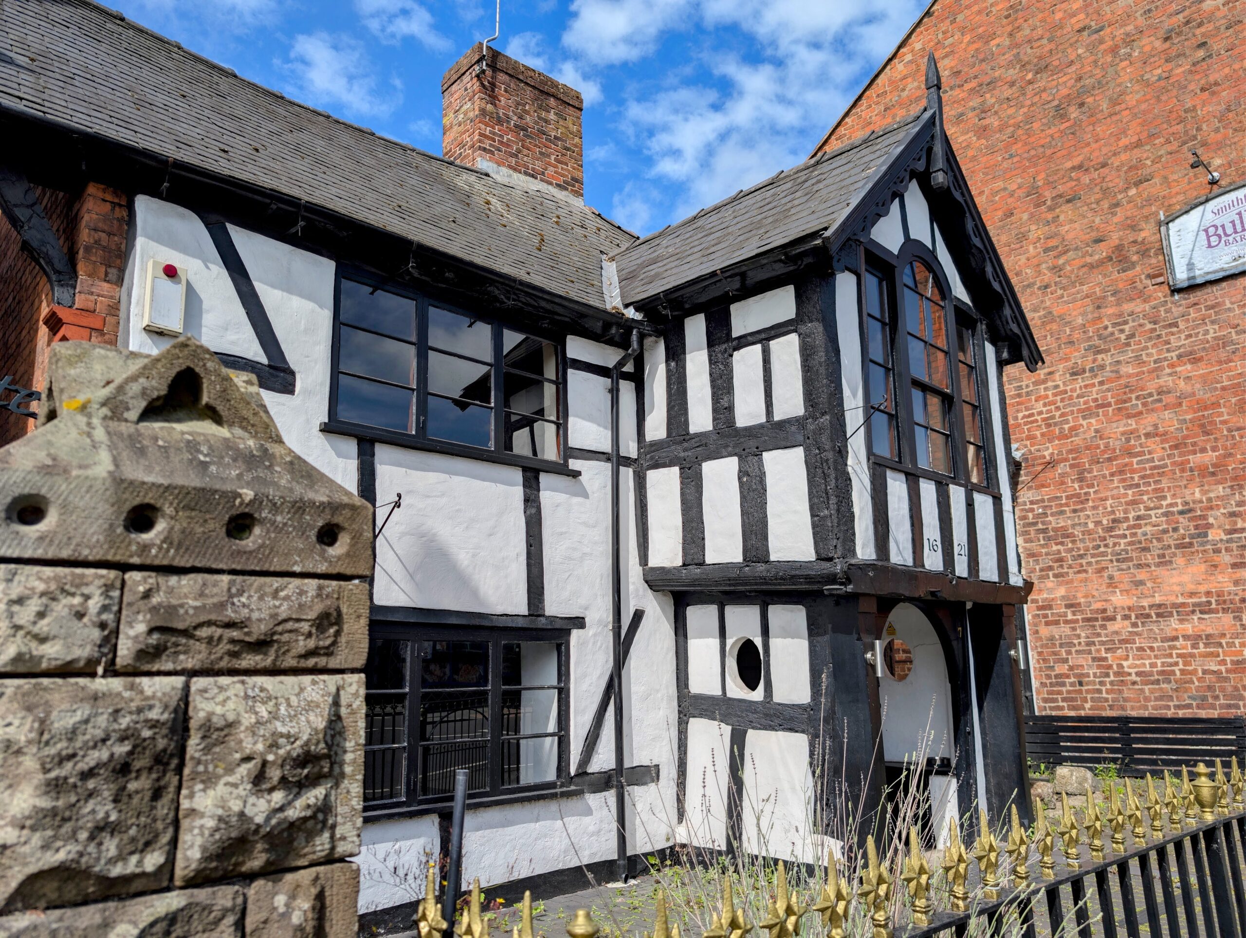 A historic, timber-framed building known as the Black Gate, featuring a classic white exterior with exposed black wooden beams. The two-story structure has multi-paned windows, a prominent gabled porch entryway with the year "1621" faintly visible on the woodwork, and a stone wall and metal railings in the foreground under a blue sky with light clouds.