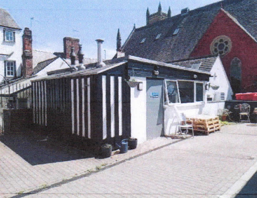 Exterior view of a commercial property's paved courtyard and outbuilding. The structure features a distinctive black and white timber-striped facade with a modern grey entrance door and large window, offering private outdoor space or main-street access potential.