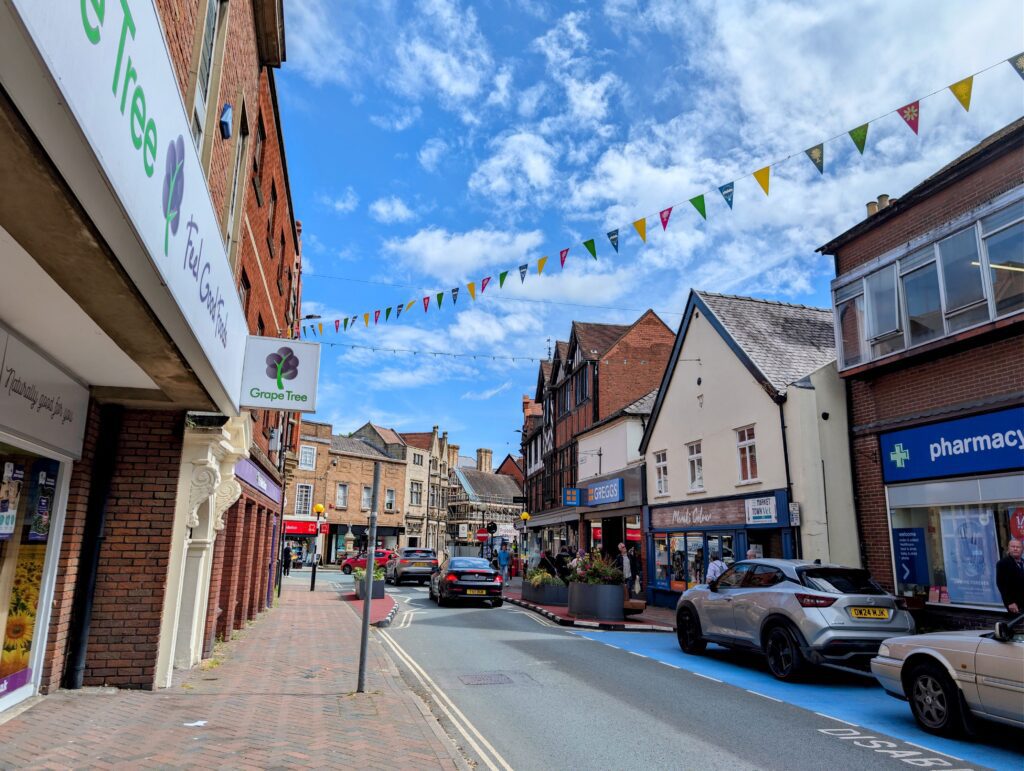A vibrant high street view of Church Street in Oswestry under a bright blue sky with scattered clouds. Colorful bunting stretches across the street above a mix of independent and high street shops, including Grape Tree, Greggs, and a local pharmacy. Several cars are parked or driving along the road, which features a designated blue-painted cycle or parking lane, surrounded by historic brick and timber-framed buildings.