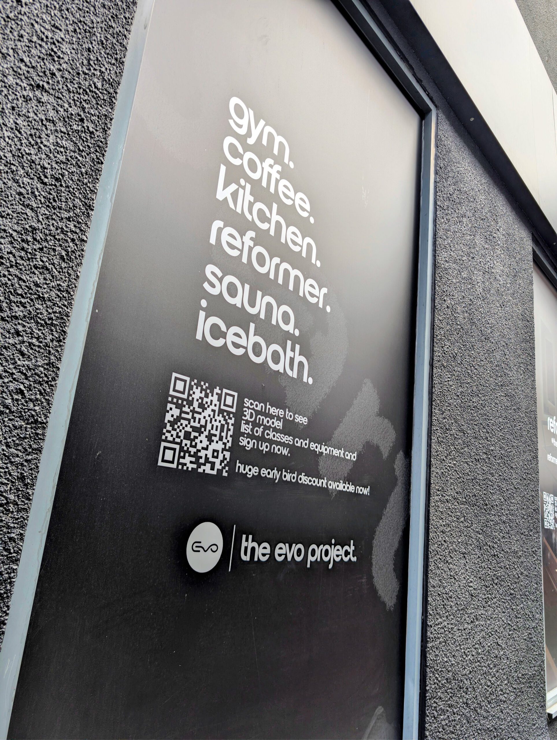 A close-up, angled shot of a black window vinyl sign on a textured grey building exterior. The white text on the sign lists "gym. coffee. kitchen. reformer. sauna. icebath." vertically in a clean, sans-serif font. Below the list is a prominent QR code with text reading "scan here to see 3D model list of classes and equipment and sign up now. huge early bird discount available now!". The bottom of the sign features the business logo and name, "the evo project.".